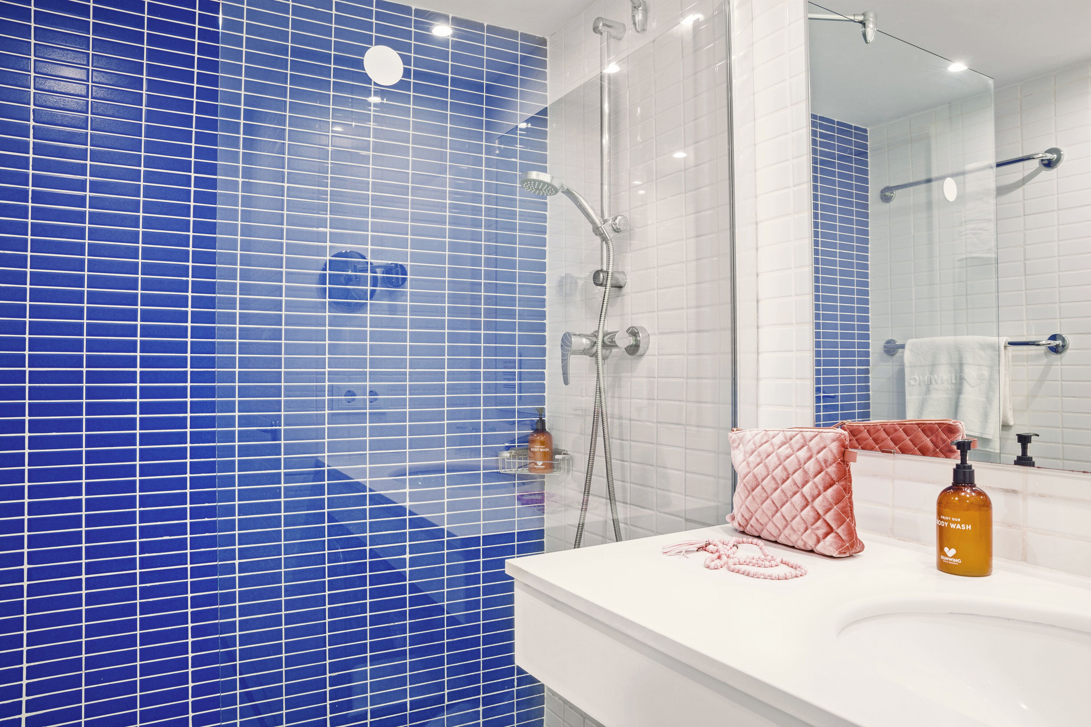 Ein Badezimmer mit blauen Fliesen, einem weißen Waschbecken, einem rosafarbenen Handtuch und einer Flasche Duschgel.