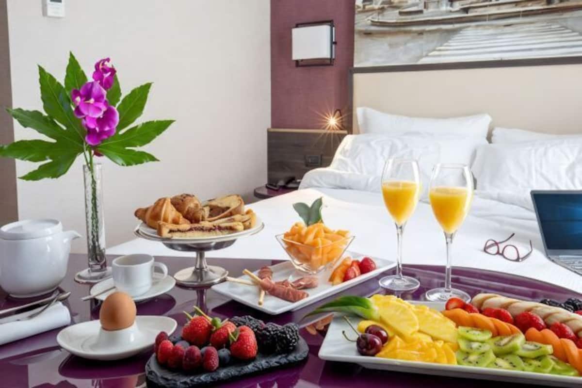 A breakfast setup with croissants, fruit, and juice glasses on a table, with a bed and a laptop in the background.