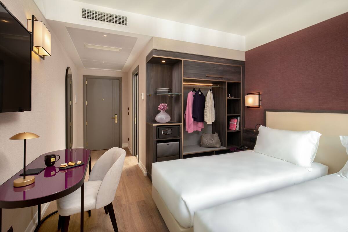 A hotel room with a bed, a desk with a lamp, a chair, a wardrobe, and a mirror.