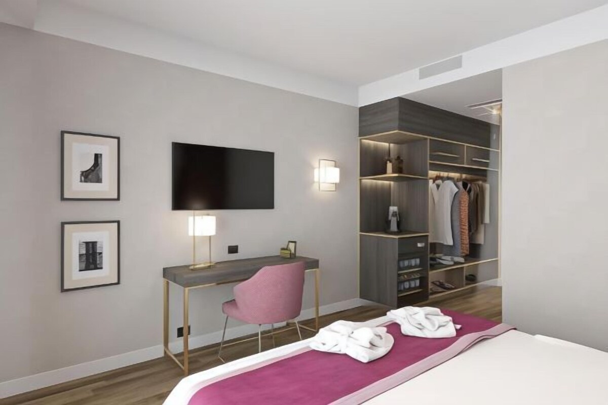 Ein modernes Hotelzimmer mit Bett, Schreibtisch, Stuhl, Fernseher und begehbarem Kleiderschrank.