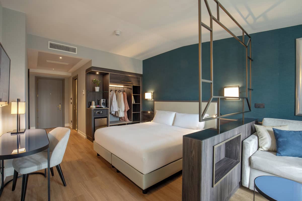 A modern hotel room with a large bed, a desk with a chair, a wardrobe, and a sofa.