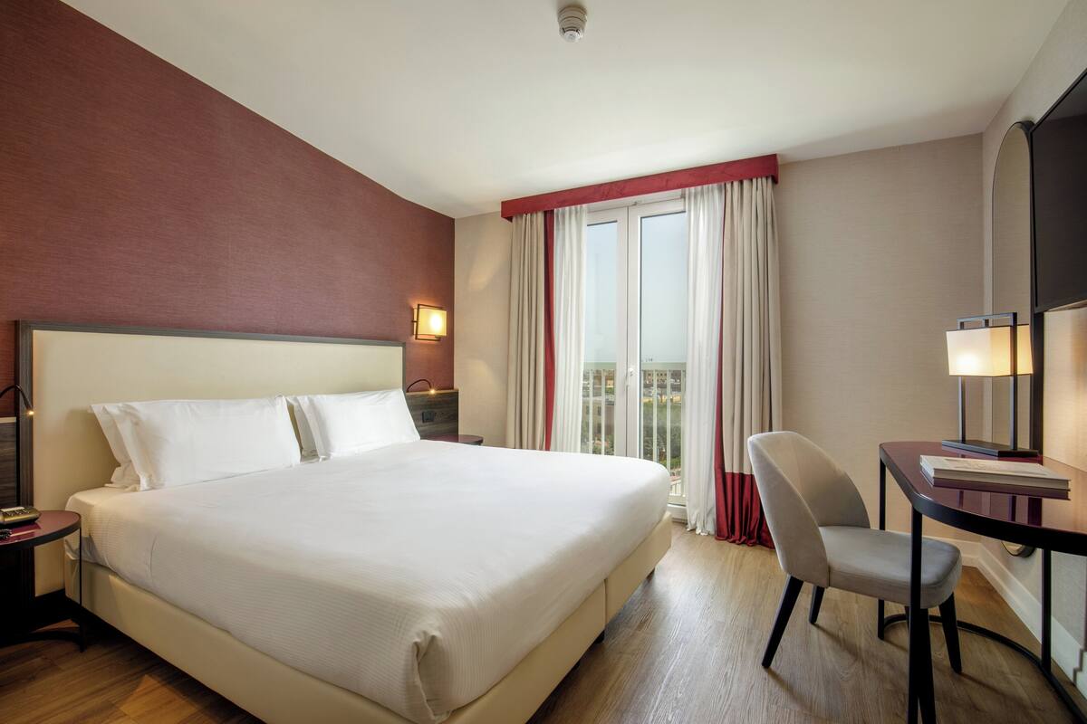 A hotel room with a large bed, a desk with a chair, a TV, and a window with curtains.