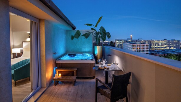 Un balcón con jacuzzi, una mesa con vasos y aperitivos, y vista a la ciudad al anochecer.