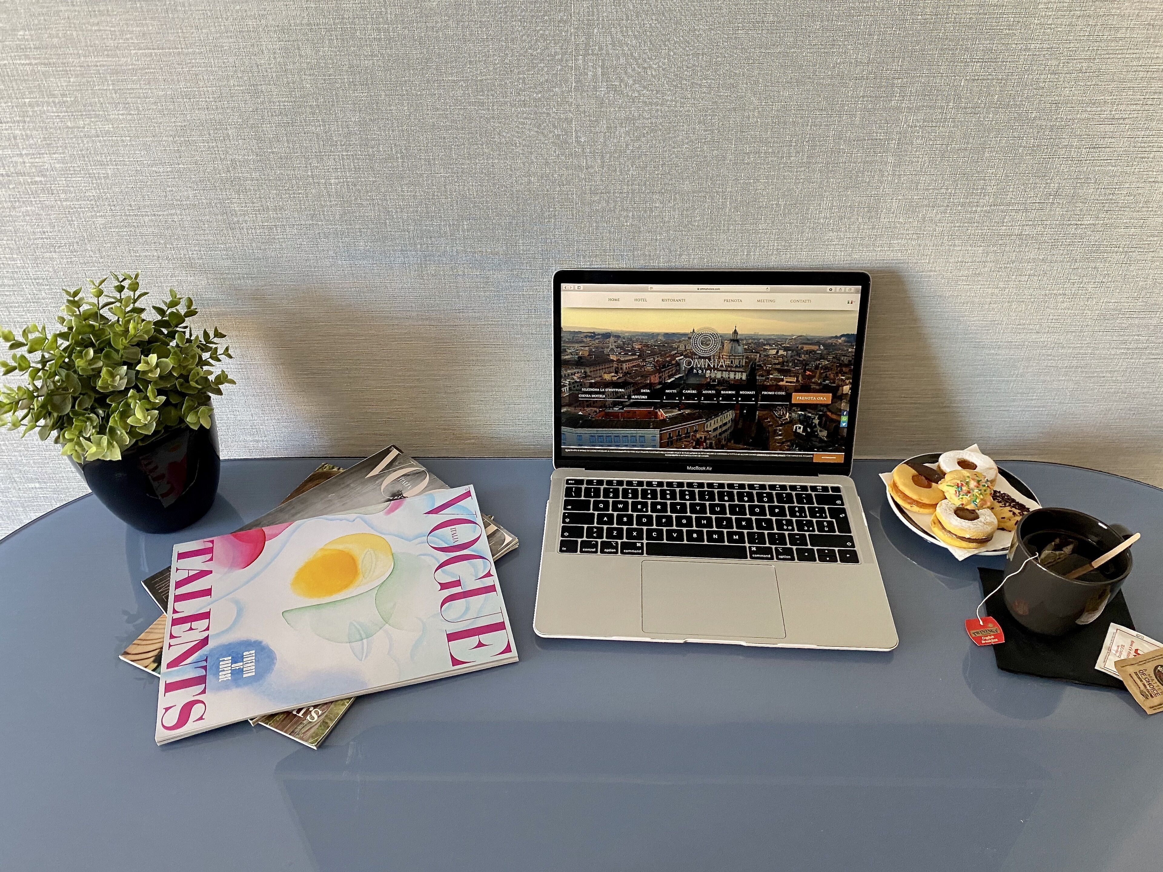 Una computadora portátil muestra un paisaje urbano, una planta, una revista y un plato de comida sobre una mesa.