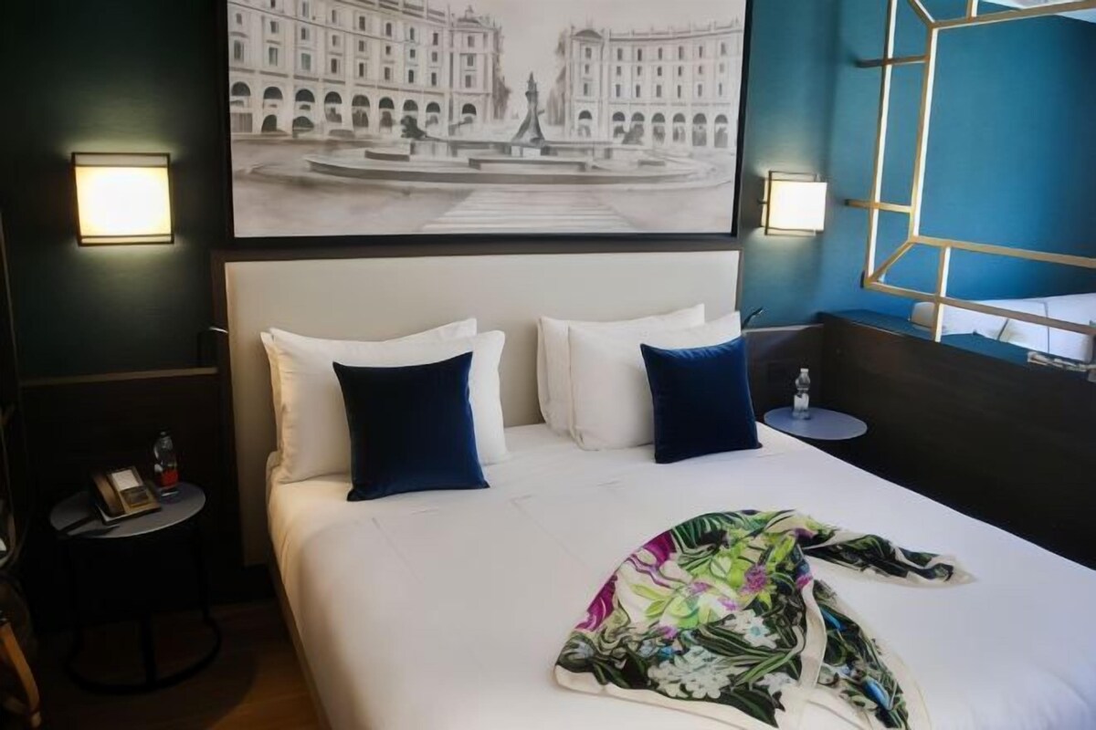 Ein Hotelzimmer mit einem großen Bett, zwei Kissen, einem Nachttisch, einem gerahmten Bild an der Wand und einem Fenster mit geometrischem Rahmen.