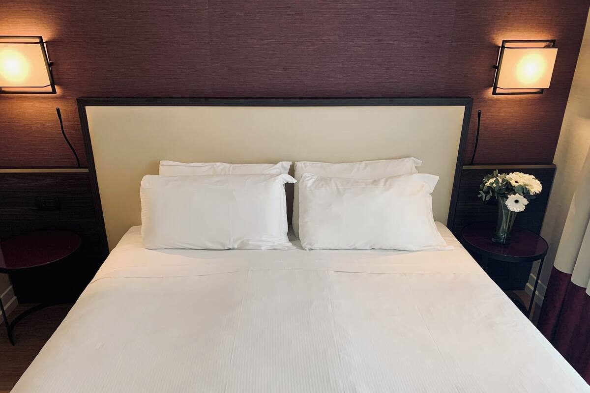 A hotel room with a large bed, two bedside lamps, a small table with a chair, and a vase with flowers on a nightstand.