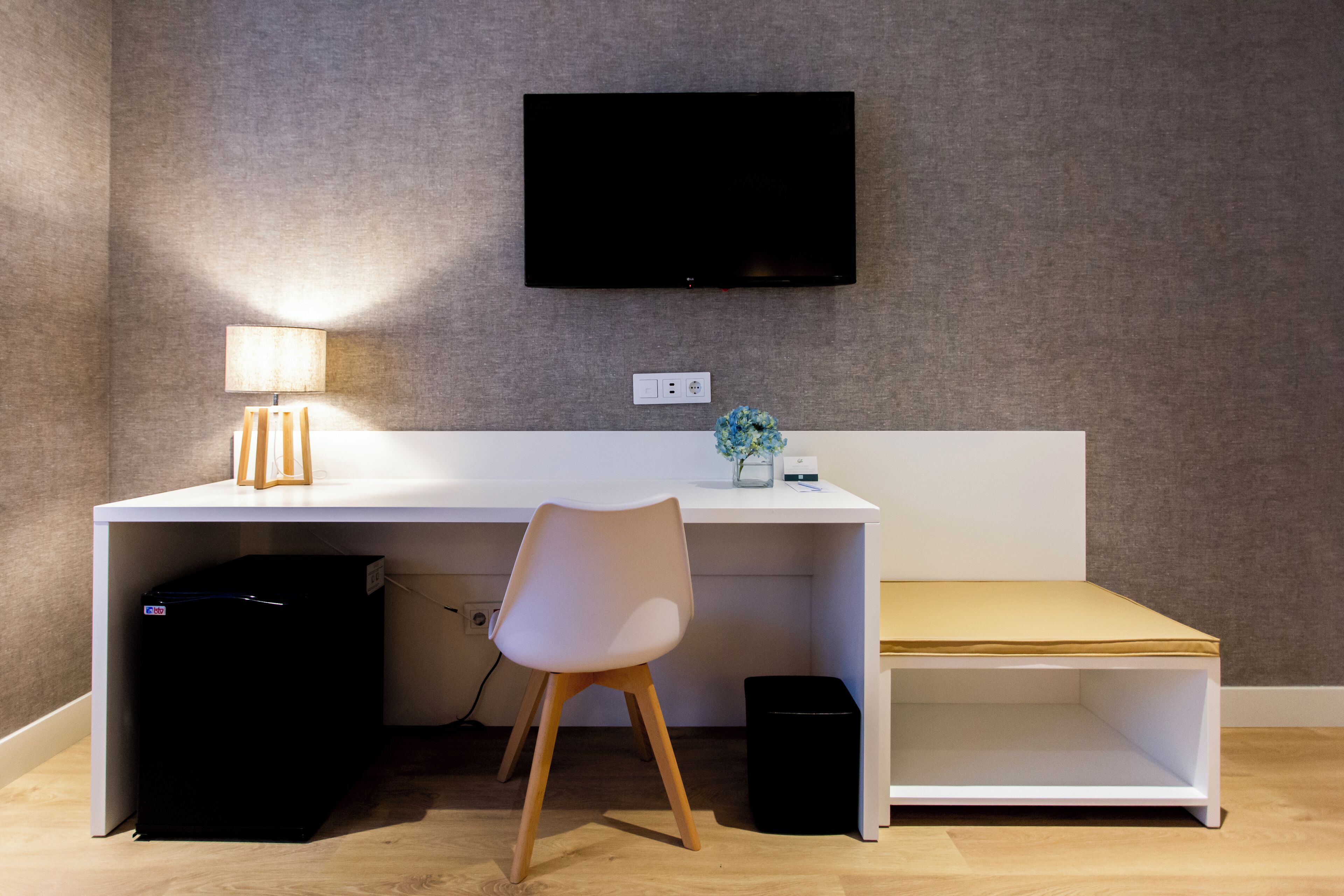 Un espacio de trabajo moderno con un televisor de pantalla plana montado en la pared, un escritorio con una silla, una lámpara, un jarrón pequeño con flores y un cubo de basura.