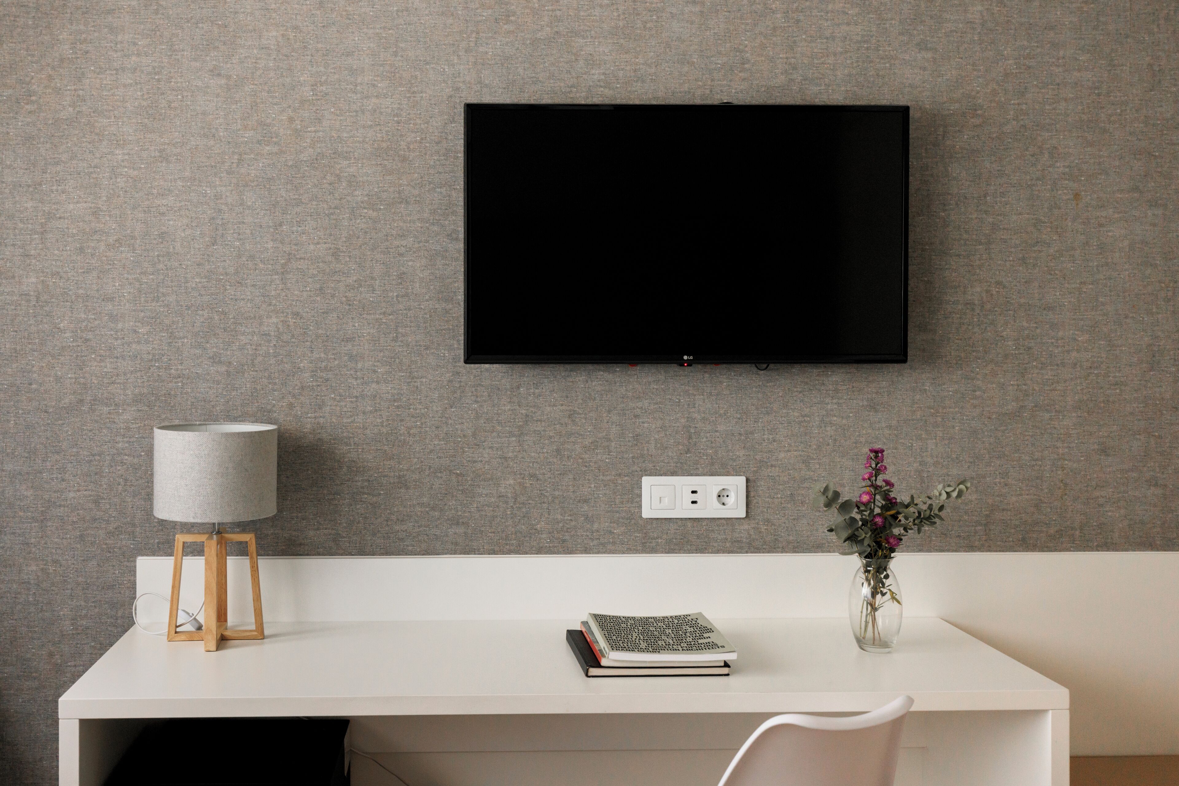 A modern room with a white desk, a mounted TV, a lamp, a vase with flowers, and a book.