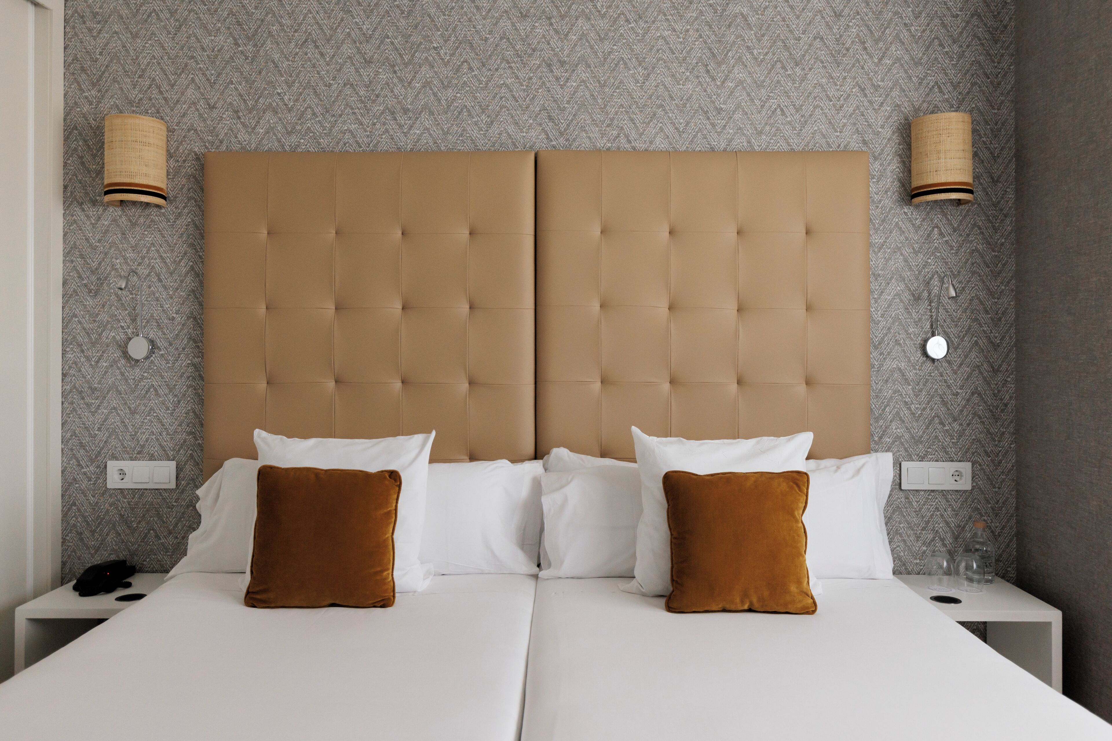Cama de casal com cabeceira estofada, roupa de cama branca e duas almofadas castanhas. Há duas mesas de cabeceira com abajures e uma tomada na parede.