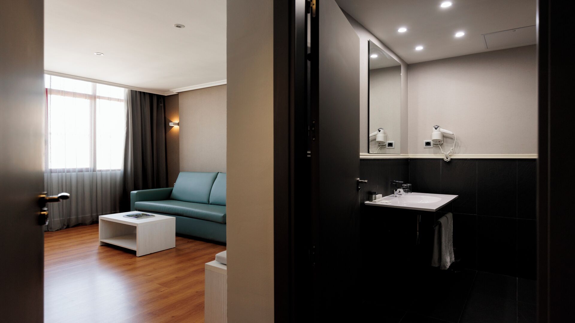 Une chambre d’hôtel moderne, dotée d’un canapé, d’une petite table blanche, d’un grand miroir et d’une salle de bain avec un lavabo et un porte-serviettes.