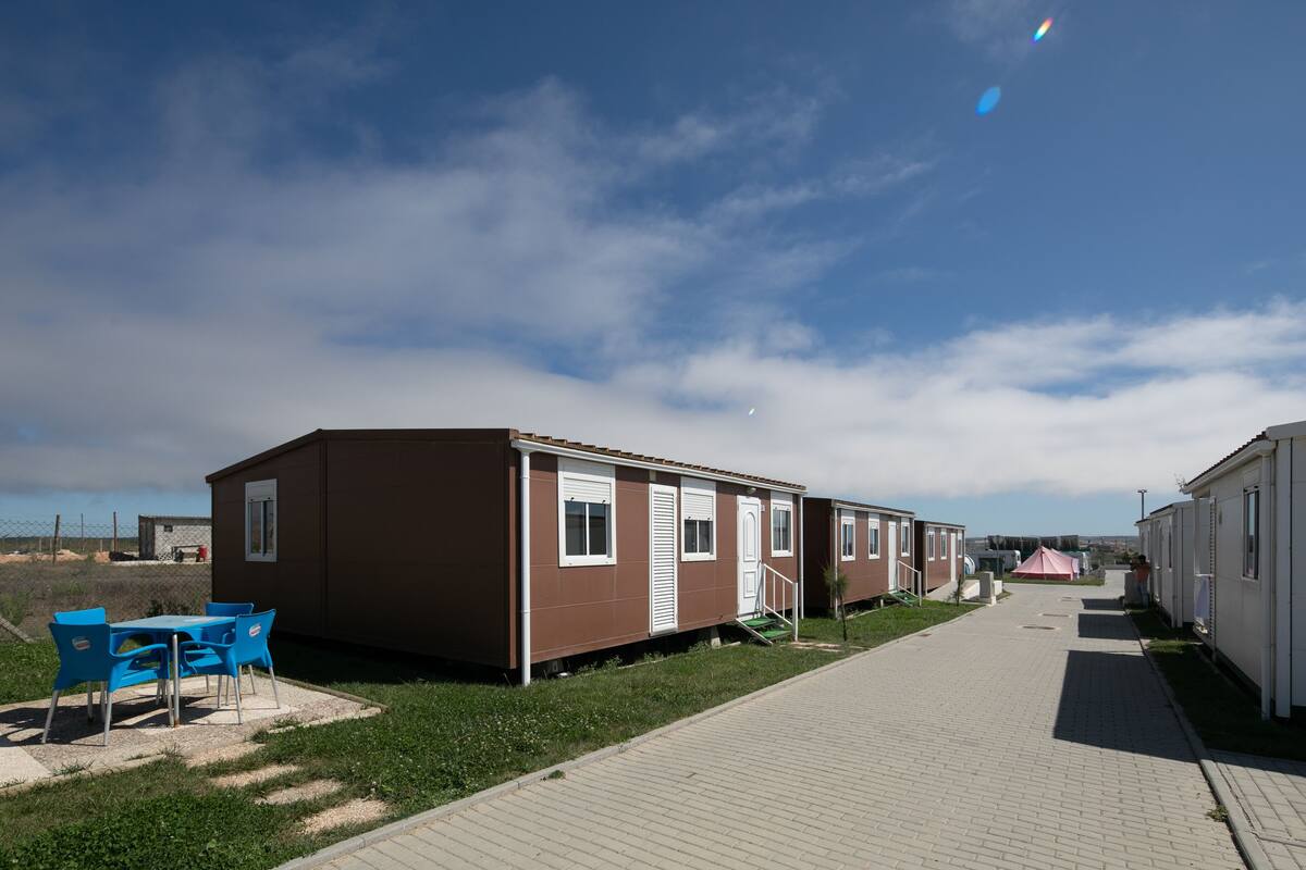 A row of mobile homes with a paved walkway, a small table with chairs, and a grassy area.