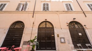 Une façade d’immeuble avec trois grandes portes noires, deux fenêtres avec volets et un pot de fleurs au premier plan.