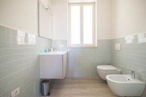 A bathroom with a toilet, bidet, and sink, light blue tiles, and a window.