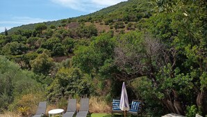 Zona de piscina con sillones de descanso y una mesa, rodeada de vegetación y colinas al fondo.