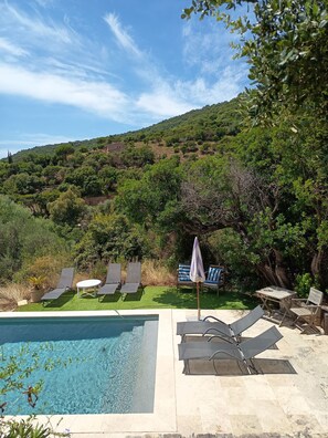 Zona de piscina con sillones de descanso y una mesa, rodeada de vegetación y colinas al fondo.
