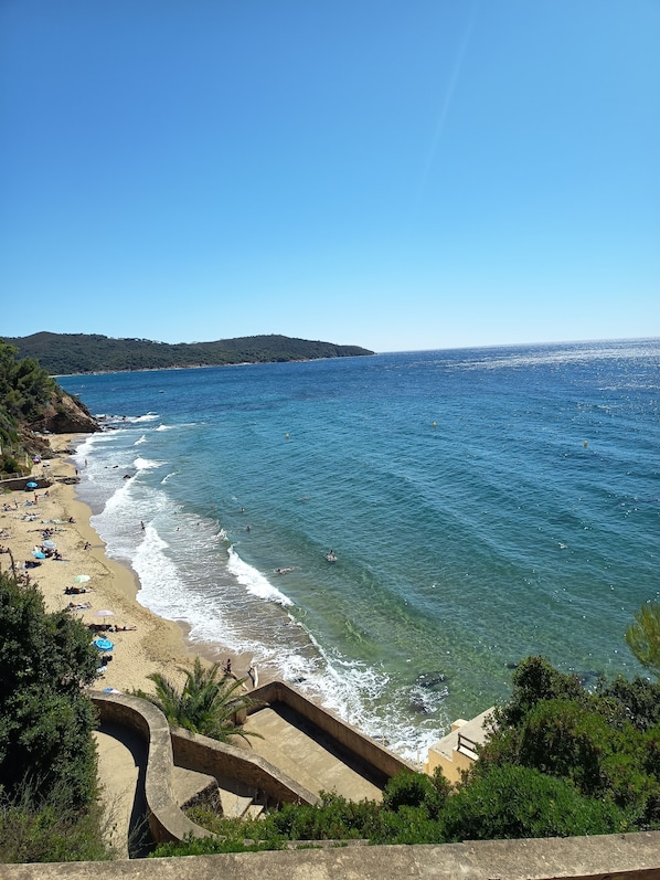 A coastal beach with clear blue waters, a sandy shore, and a hilly landscape in the background.