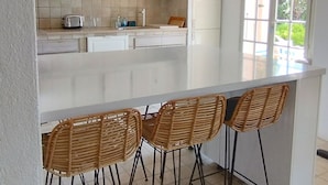 Una cocina con armarios blancos, encimera blanca y tres taburetes de mimbre.