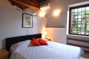 A bedroom with a bed, a nightstand, a lamp, a framed picture, an air conditioning unit, and a window with a view of trees.