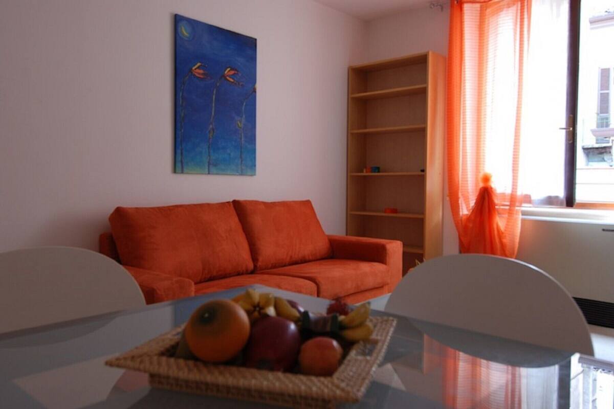 Uma sala de estar com um sofá vermelho, uma cesta de frutas sobre uma mesa de vidro, uma estante de livros e um quadro na parede.