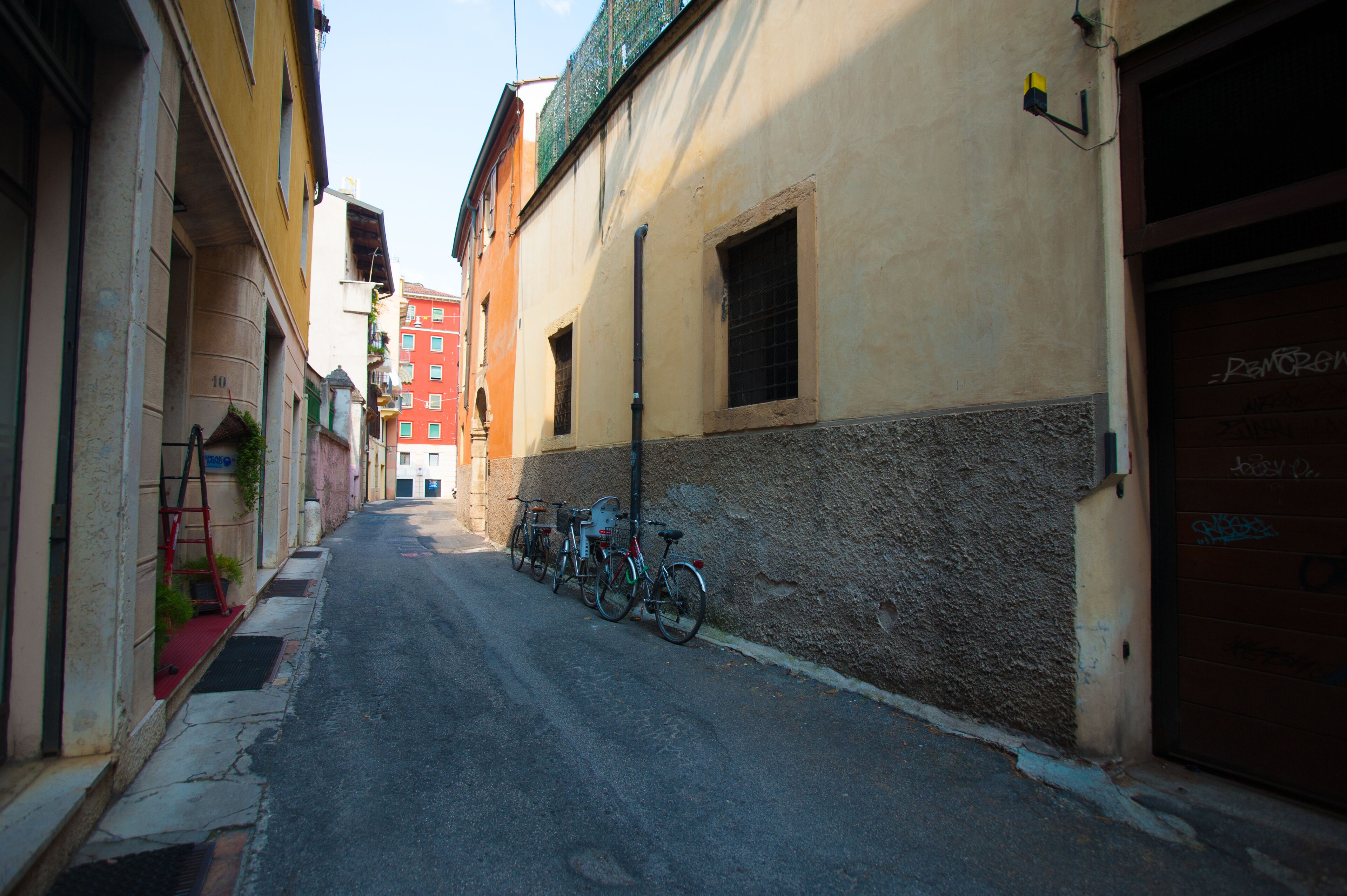 A narrow street with buildings on either side, bicycles parked along the curb, and a red building in the background.