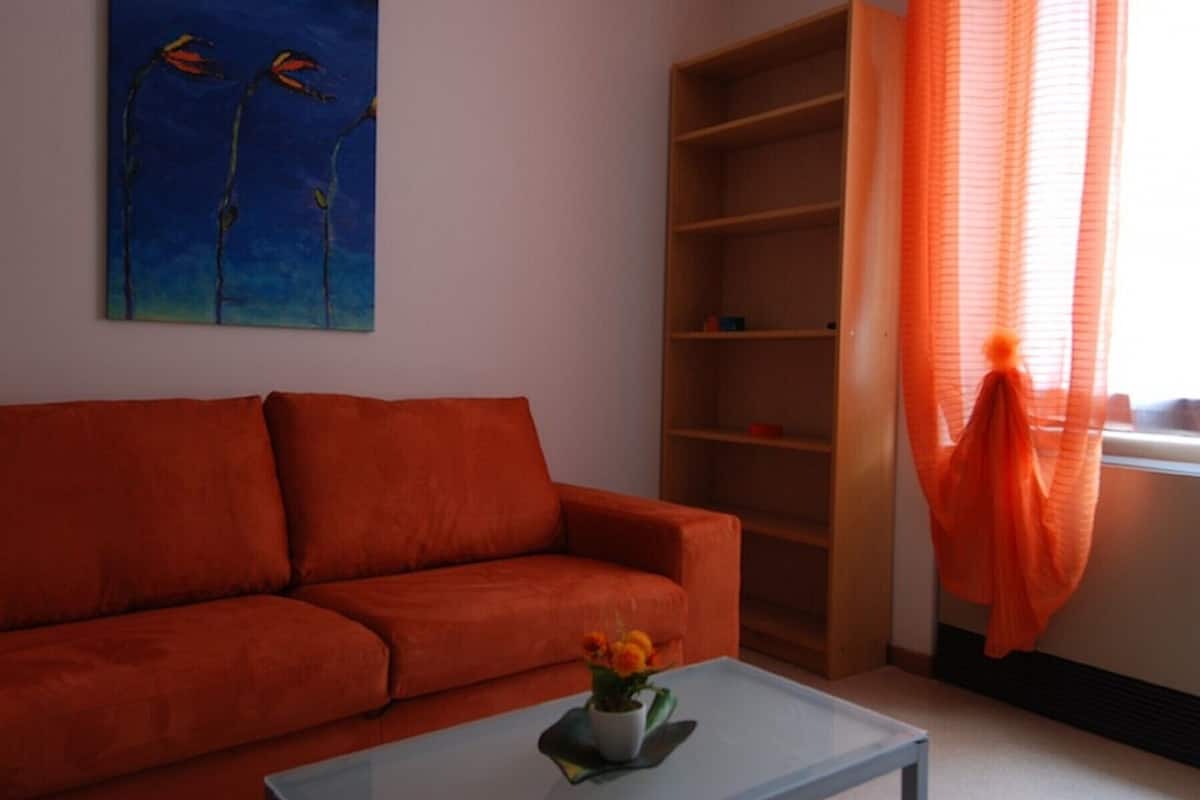 Uma sala de estar com um sofá vermelho, uma mesa de centro de vidro com um vaso de flores, uma estante e uma janela com cortinas laranja.