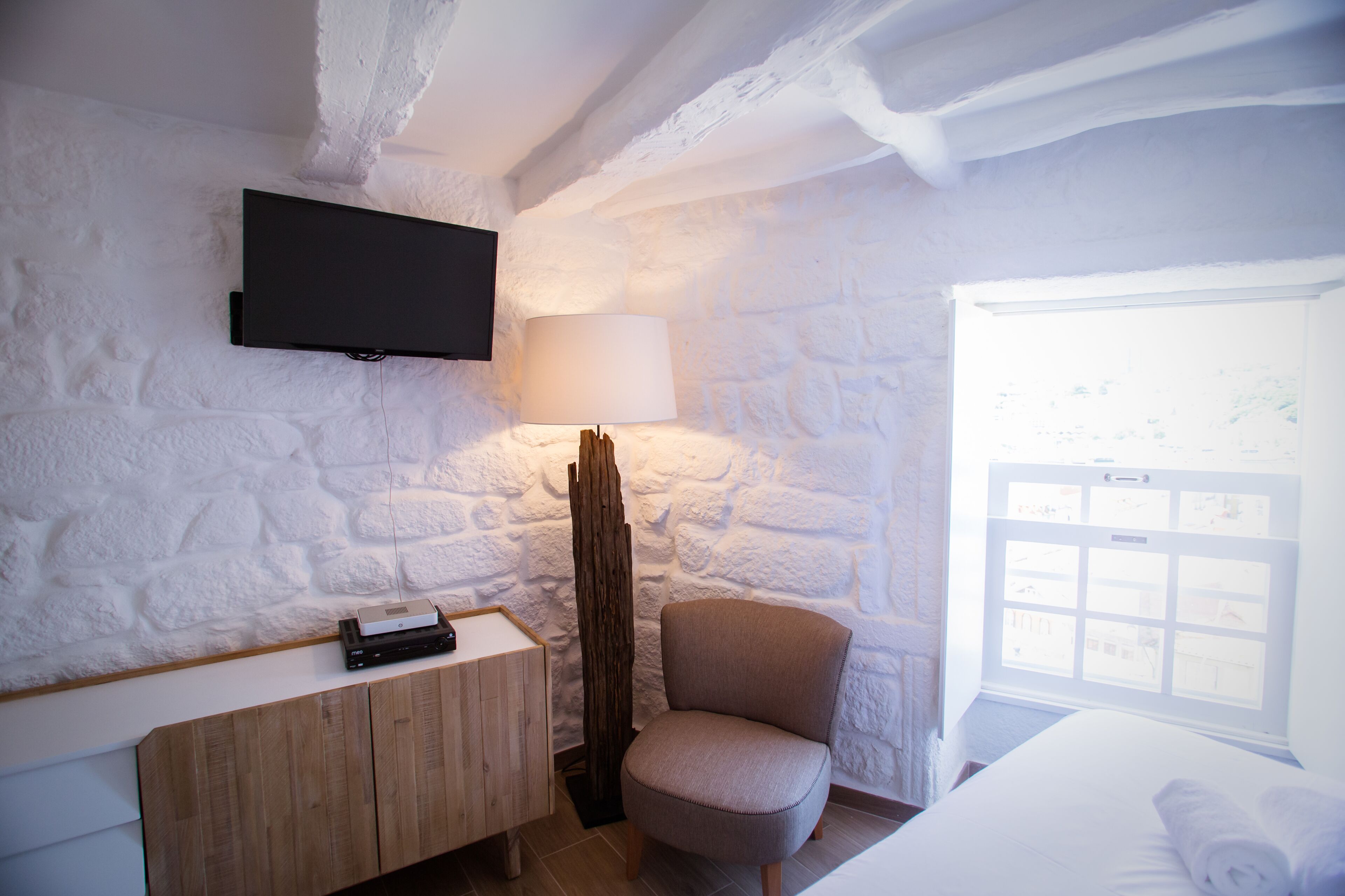 A room with a stone wall, a mounted TV, a wooden cabinet, a chair, a lamp, and a window with a view.
