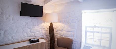 A room with a stone wall, a mounted TV, a wooden cabinet, a chair, a lamp, and a window with a view.