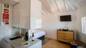 Quarto de hotel com uma cama, uma escrivaninha, televisão, espelho, pia e janela.