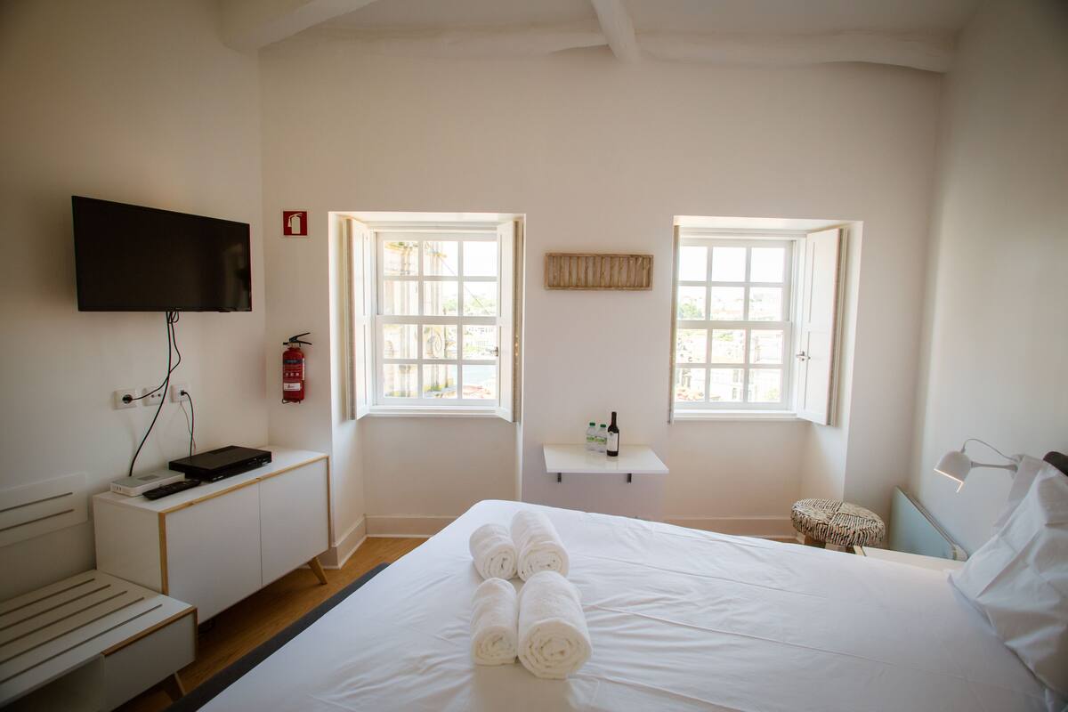 Ein Hotelzimmer mit einem Bett, einem an der Wand befestigten Fernseher, einem kleinen Tisch mit einer Flasche sowie zwei Fenstern.