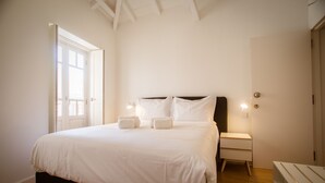 Cama bem arrumada com lençóis brancos, mesa de cabeceira com abajur e um banco de madeira.