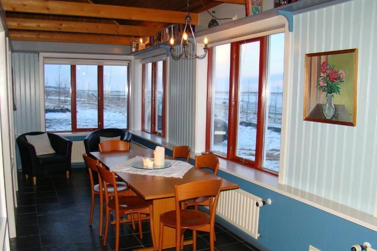 Uma sala de jantar com uma mesa posta para quatro pessoas, rodeada por cadeiras, grandes janelas com vista para uma paisagem coberta de neve, um candelabro e um quadro na parede.