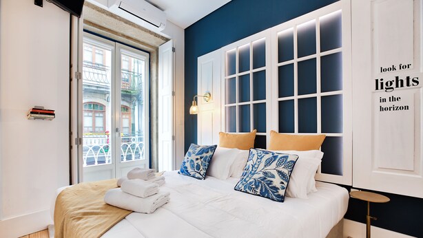 A neatly made bed with white linens and blue patterned pillows, a bedside table with a lamp, and a view of a building through a window with white shutters. - Janelas de São Bento by Porto City Hosts (Porto)