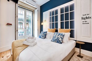 Un letto rifatto con lenzuola bianche e cuscini azzurri a motivi decorativi, un comodino con una lampada e vista su un edificio attraverso una finestra con persiane bianche.