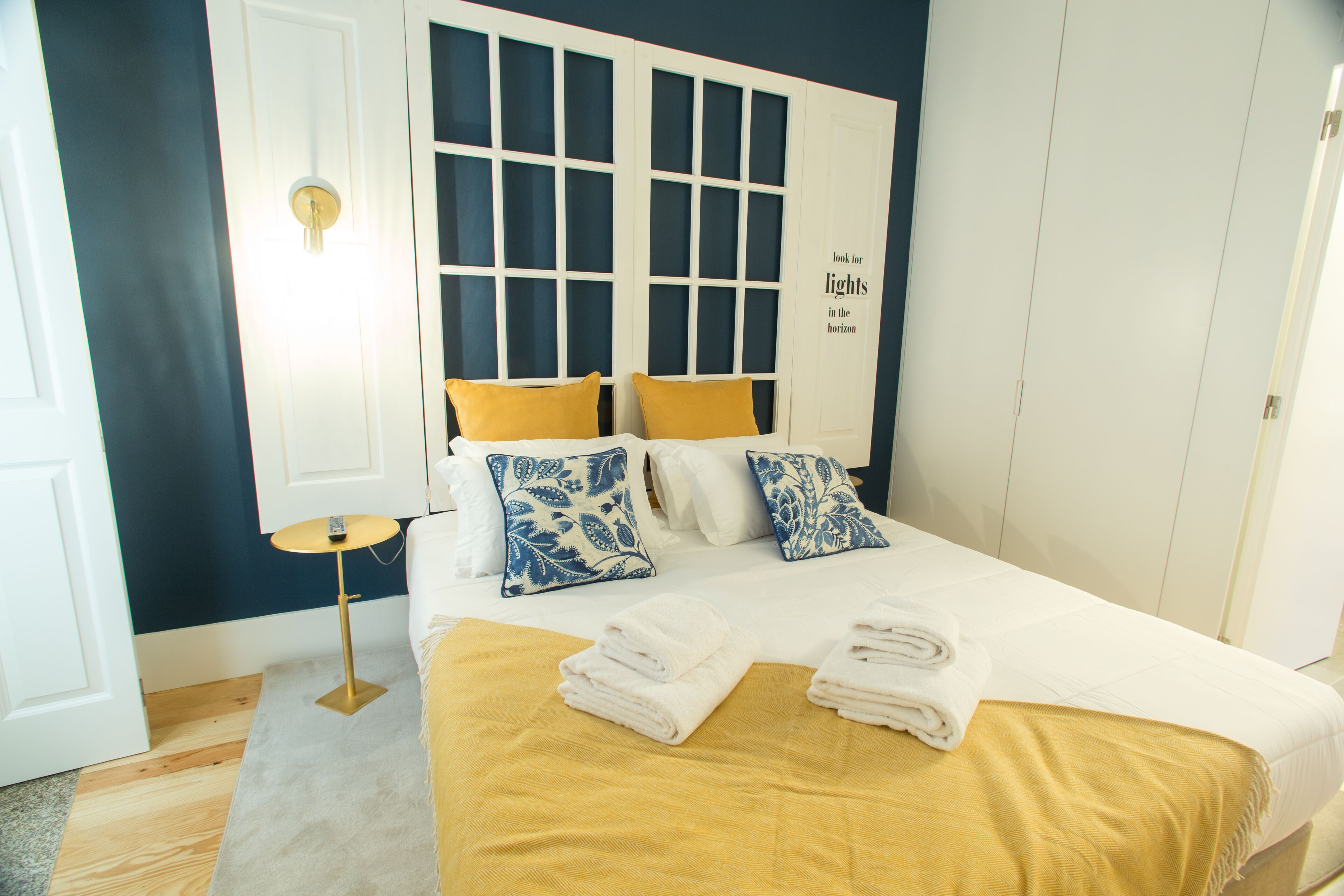 A neatly made bed with a yellow bedspread and two pillows, a bedside table with a lamp, and a wall with a window and a sign.