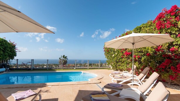 Zona junto a la piscina con sillones de descanso y sombrillas, con vista a un cielo azul despejado y al océano.