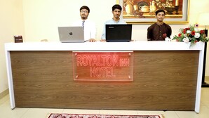 A reception desk at the Royalton Inn Hotel with three individuals standing behind it, laptops on the desk, and a floral arrangement to the right. - Royalton Inn Hotel (Lahore)
