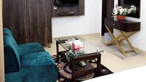 A modern living room with a teal sofa, a glass coffee table, a TV mounted on the wall, and a wooden wardrobe. - Royalton Inn Hotel (Lahore)