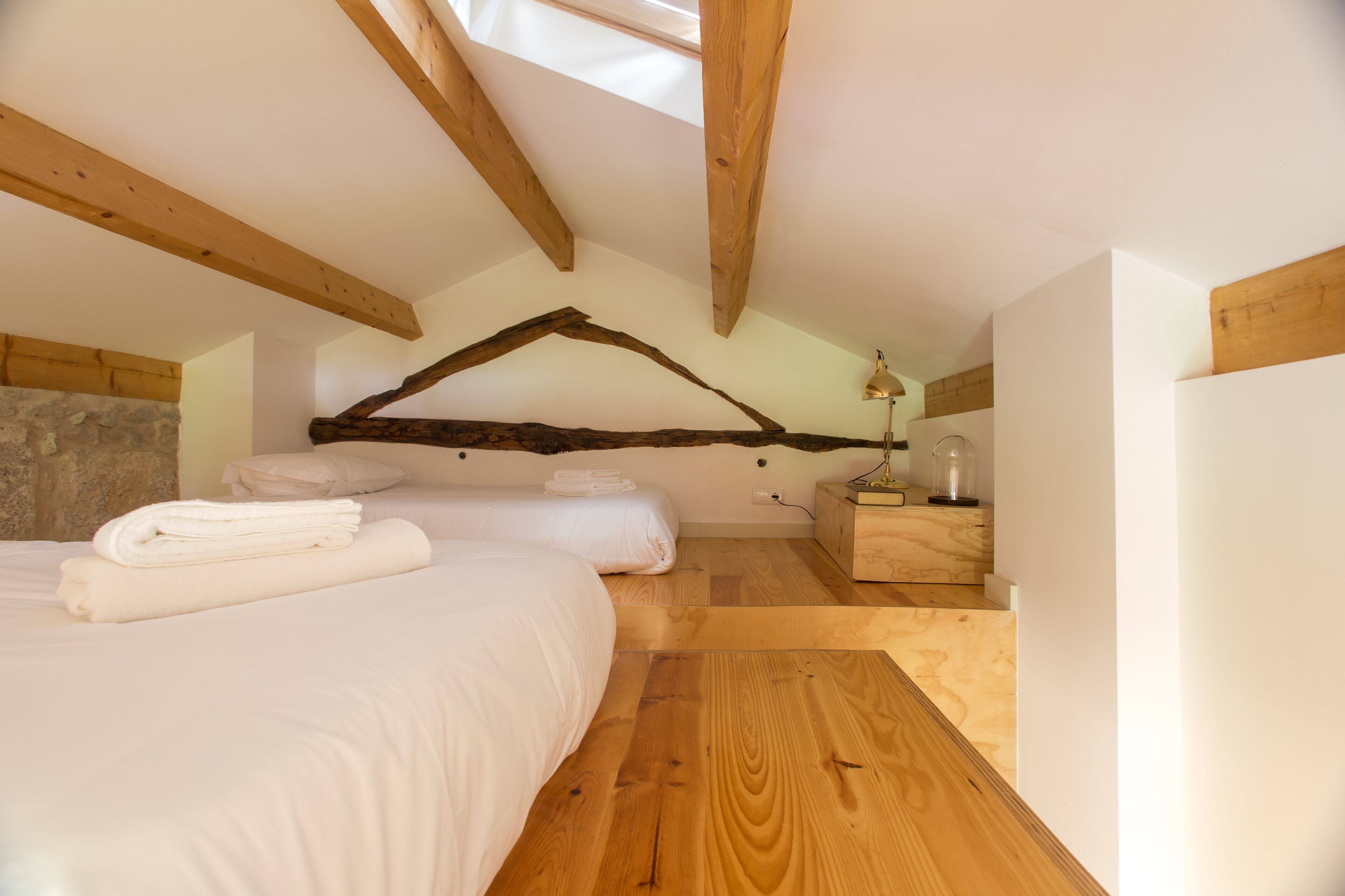 A bedroom with a sloped ceiling, wooden beams, a bed with white linens, a nightstand with a lamp, and a stone wall.
