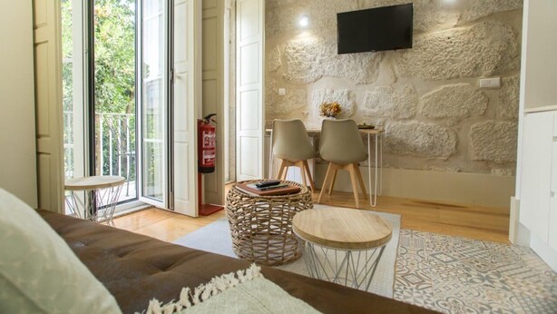 A modern living room with a stone wall, a flat-screen TV, a sofa, two chairs, a small round table, and a wicker ottoman. - Picasso Suites Porto by Porto City Hosts (Porto)
