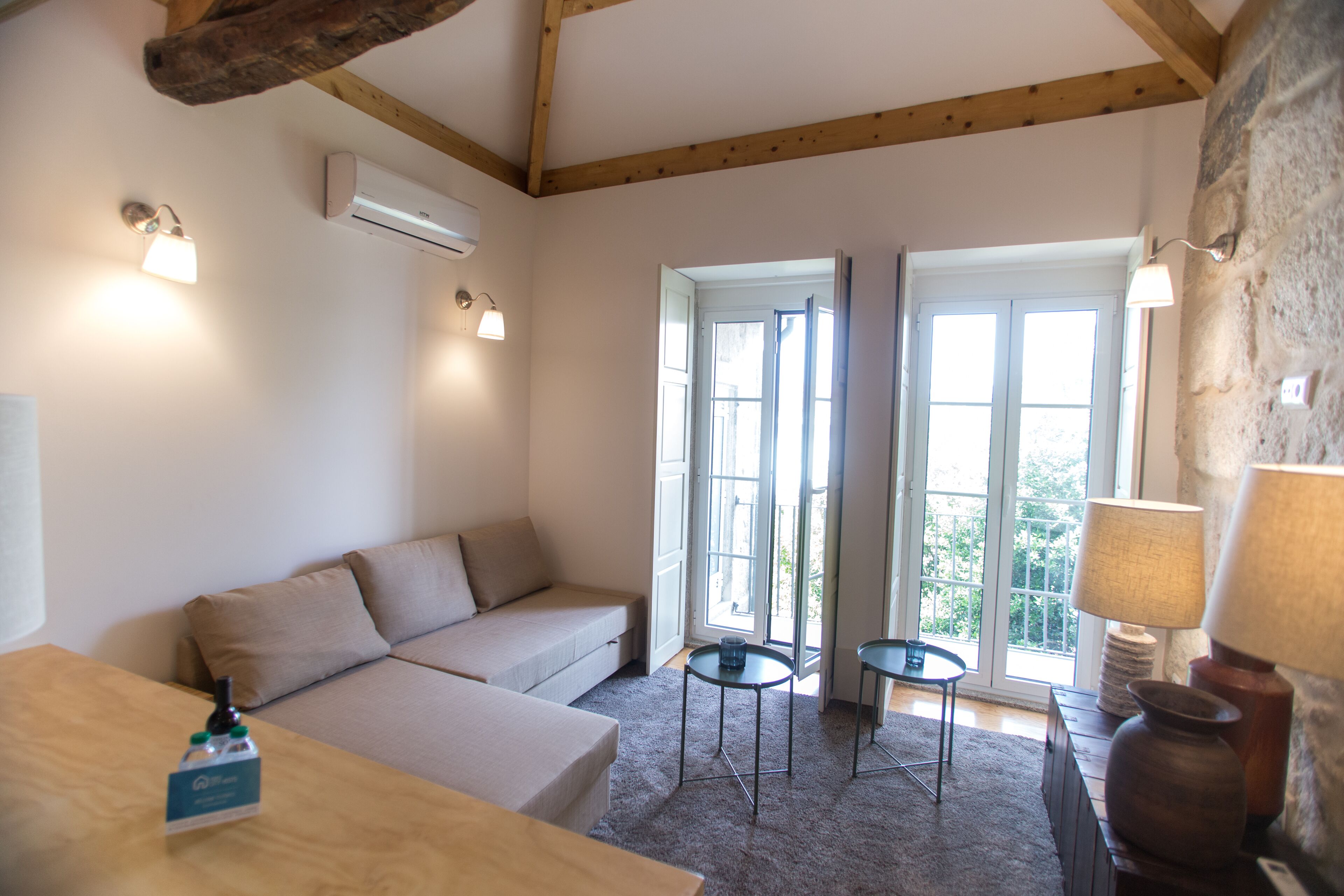 A cozy room with a sofa, a small table, and a lamp. There are two windows with curtains, an air conditioning unit, and a stone wall.