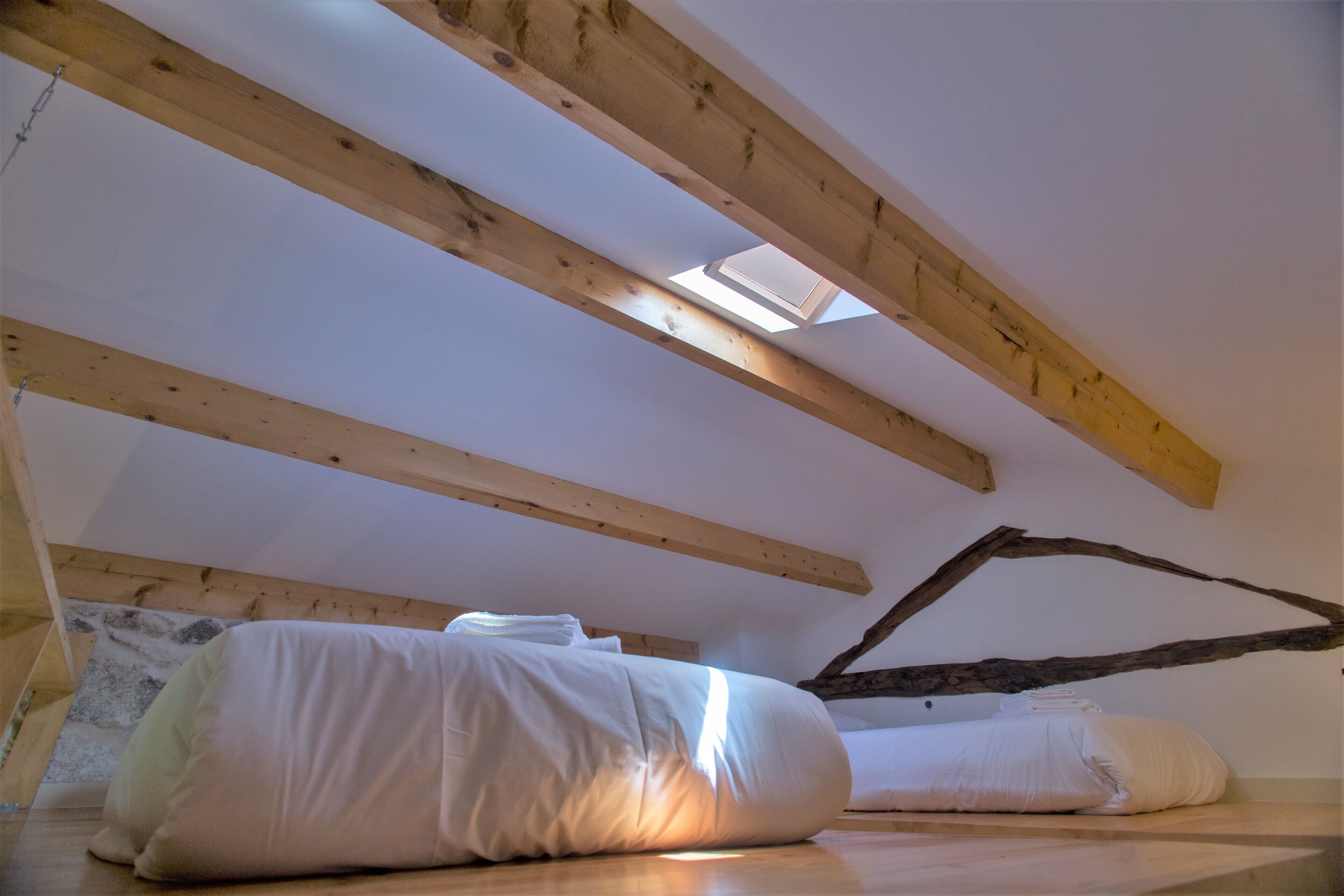 A room with a sloped ceiling, wooden beams, a skylight, and a bed with white bedding.