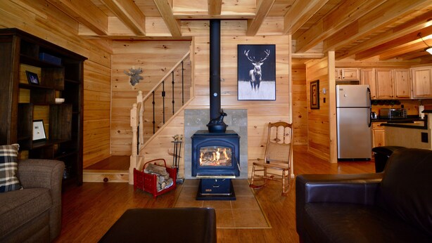 A log cabin interior with a fireplace, wooden walls, a kitchen area, and a staircase.