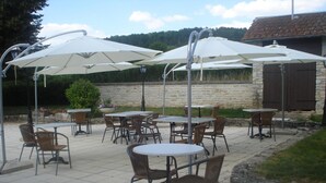 Een terras buiten met tafels en stoelen, grote witte parasols en een stenen muur op de achtergrond.