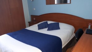 Een net opgemaakt bed met blauw beddengoed, een houten hoofdbord en een klein nachtkastje met een bloevas.