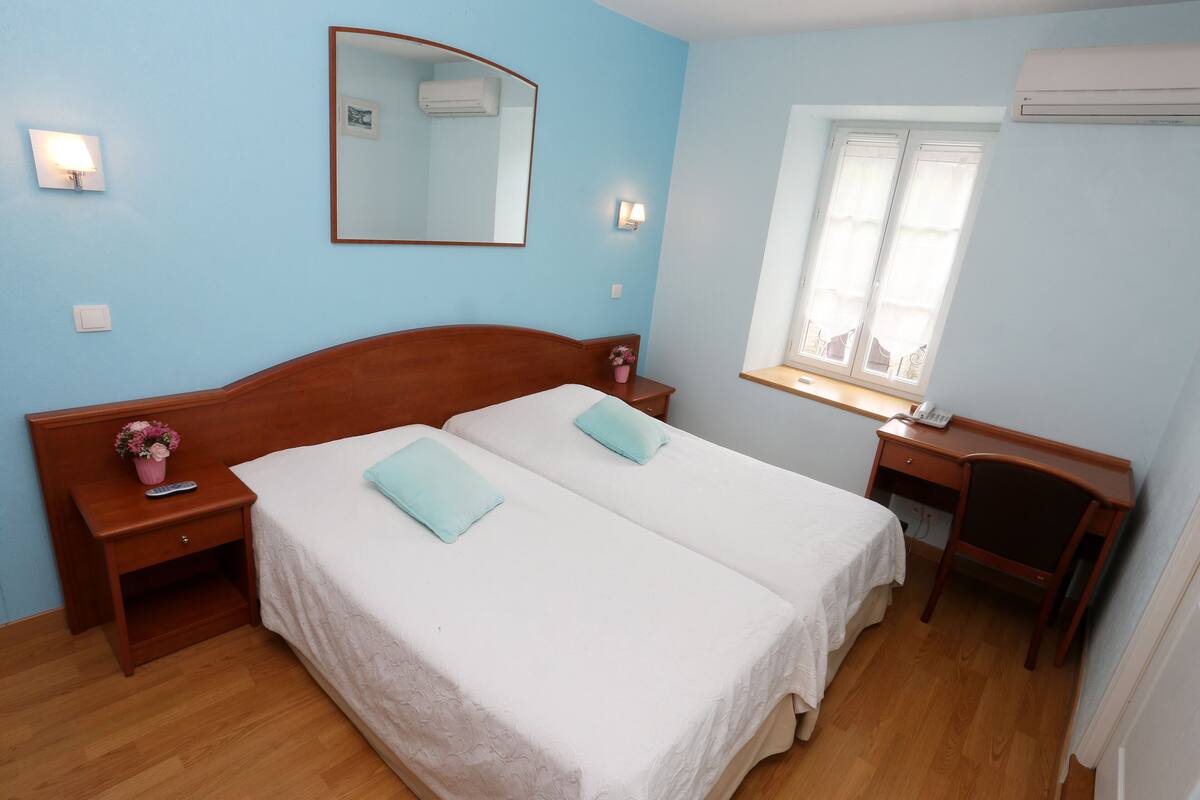 Ein Schlafzimmer mit zwei Betten, einem Schreibtisch, einem Stuhl, einem Fenster mit Vorhängen und einem an der Wand befestigten Spiegel.
