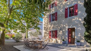 A courtyard with a wooden table and chairs, surrounded by a building with red shutters and a door. - La Maison Vigneronne (Trebes)