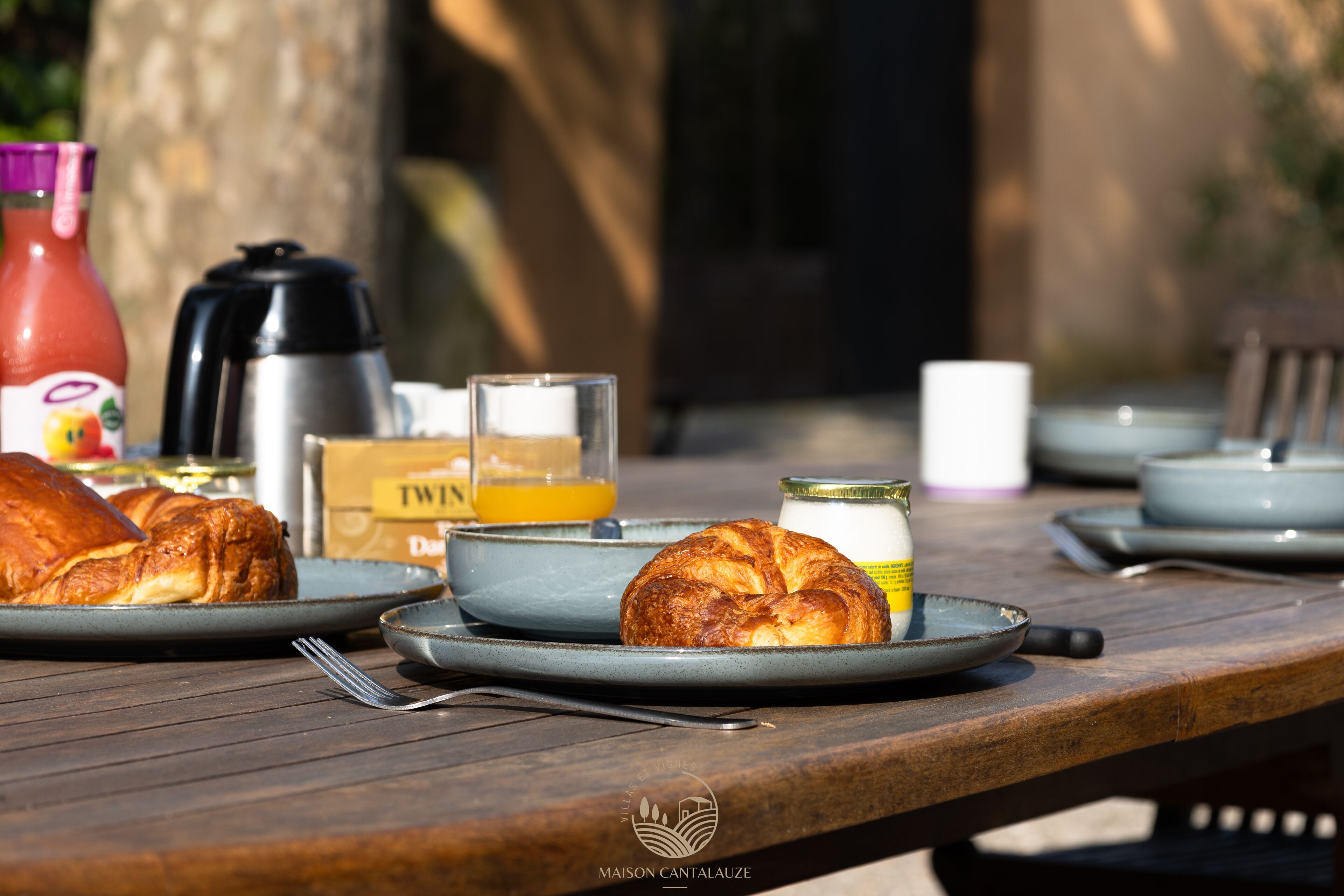 A breakfast table with croissants, a bottle of orange juice, a thermos, and a coffee maker.