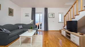 En stue med en grå sofa, et hvidt sofabord, et fjernsyn på et træstel og udsigt gennem en glasdør.