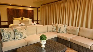 A modern living room with a large beige sectional sofa, a small round table with a potted plant, and a bed with matching pillows and a headboard.
