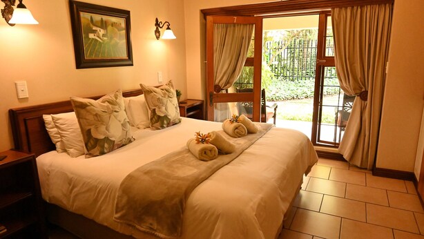 A hotel room with a large bed, a painting on the wall, a nightstand, a lamp, and a view of the garden through the door. - Lamor Guest House (Middelburg)